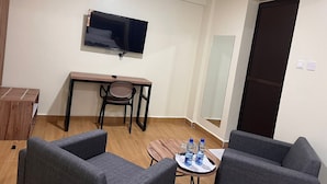 Ein kleiner, moderner Raum mit einem Flachbildfernseher an der Wand, einem Holztisch mit Stuhl, zwei grauen Sofas und einem kleinen Tisch mit Wasserflaschen.