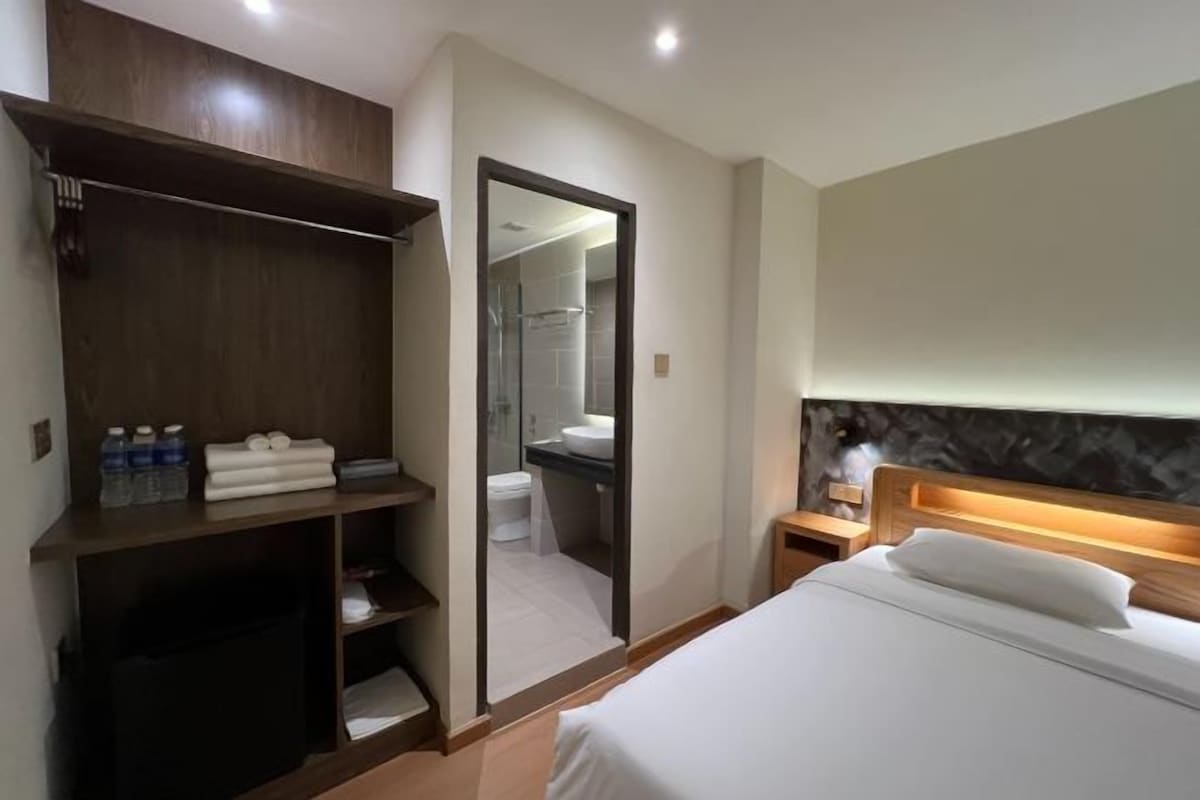 A hotel room with a bed, a wooden wardrobe, a mirror, a nightstand, and a bathroom visible through an open door.