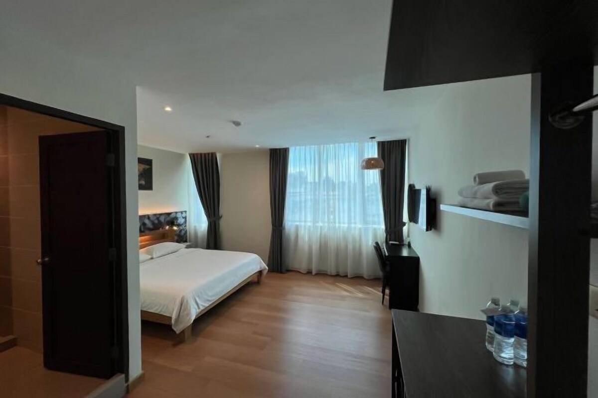 A hotel room with a bed, a desk, a chair, a window with curtains, and a shelf with towels.