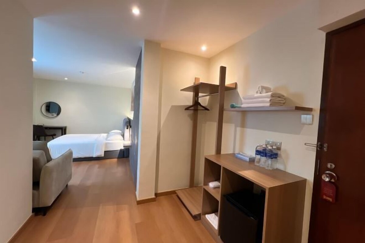 Une chambre d’hôtel équipée d’un lit, d’un bureau, d’une chaise, d’un miroir et d’une étagère où sont rangées des serviettes et des articles de toilette.