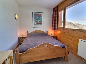 A wooden bed with a heart-shaped cutout, two bedside tables with lamps, a framed picture on the wall, and a window with a view of a snowy landscape.
