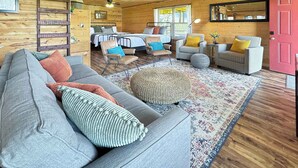 Un interior de cabaña acogedor con un sofá, sillas, un otoman redondo, una cama y techo de madera.