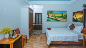 Een hotelkamer met een bed, een bureau, een stoel, een kleintje tafeltje met een plant, en kunstwerken aan de muur.