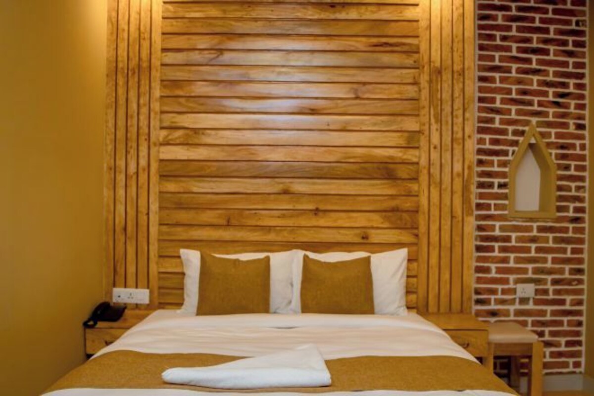 A hotel room with a wooden headboard, a brick wall, a small wooden nightstand with a phone, and a wooden chair.