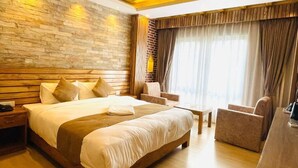 Een moderne hotelkamer met een groot bed, een houten hoofdeinde en een accentmuur van steen. Er is een zithoek met een bank en fauteuils, en een groot raam met gordijnen.