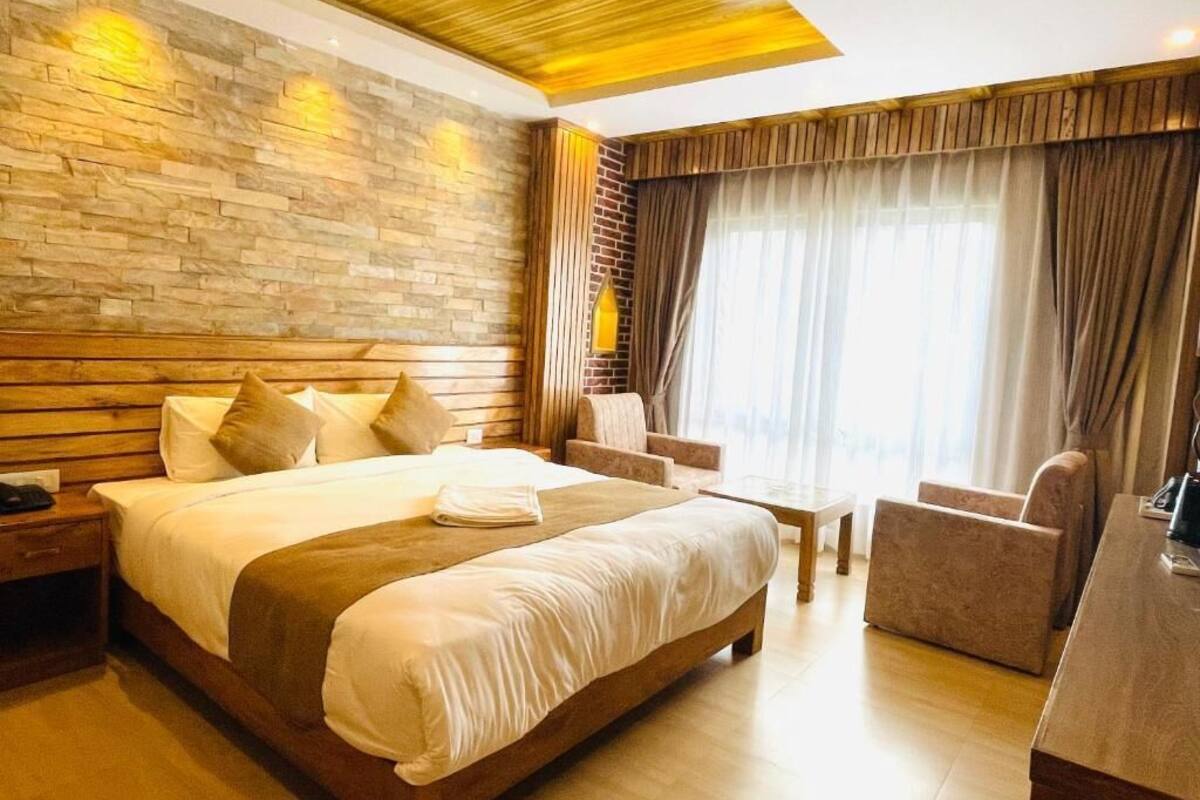A modern hotel room with a large bed, wooden headboard, and a stone accent wall. There is a seating area with a sofa and armchairs, and a large window with curtains.