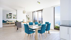 A modern dining area with a large table and blue chairs, a kitchen with white cabinets, and a view of the outdoors through large windows. - VILLA CANELA, VACATION HOME IN THE NORTH OF TENERIFE (El Sauzal)