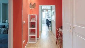 Un pasillo con piso de madera, una puerta blanca a la derecha y una pared roja con un estante blanco y un adorno.