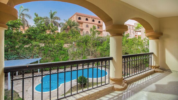A balcony with a view of a swimming pool, palm trees, and a building with a red facade. - Host Prime Paradise Villa At Rehab City (New Cairo)