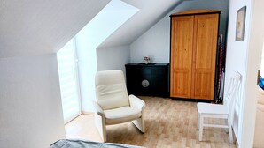 A bedroom with a sloped ceiling, a wooden wardrobe, a white chair, and a small table with a vase.