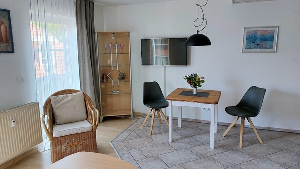 A modern living room with a wicker sofa, a wooden dining table with two chairs, a TV mounted on the wall, and a radiator.