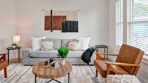 A modern living room with a grey sofa, a wooden coffee table, a brown armchair, and a wall-mounted macrame piece. - Central HTX Apt: Free Wi-Fi, A/C, Full Kitchen, Near Top Attractions. (Houston)