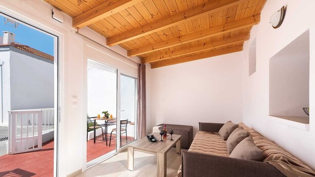 Een kamer met een houten plafond, een bank, een tafel en een glazen schuifdeur naar een buitenruimte met een tafel en stoelen.