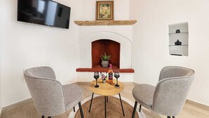 Een woonkamer met een open haard, twee grijze stoelen, een houten tafel met wijnglazen, een aan de muur bevestigde televisie en een lijst met een afbeelding boven de open haard.