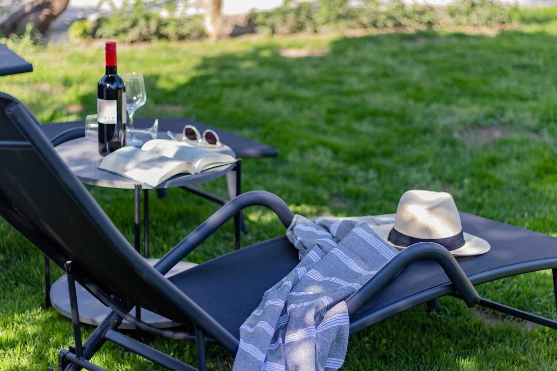 Una tumbona con sombrero, un libro, una botella de vino y gafas de sol sobre una mesa en un espacio al aire libre con césped.