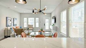 Ein geräumiges Wohnzimmer mit einem Glastisch, bequemen Sitzgelegenheiten, einem modernen Kronleuchter und großen Fenstern mit Jalousien.