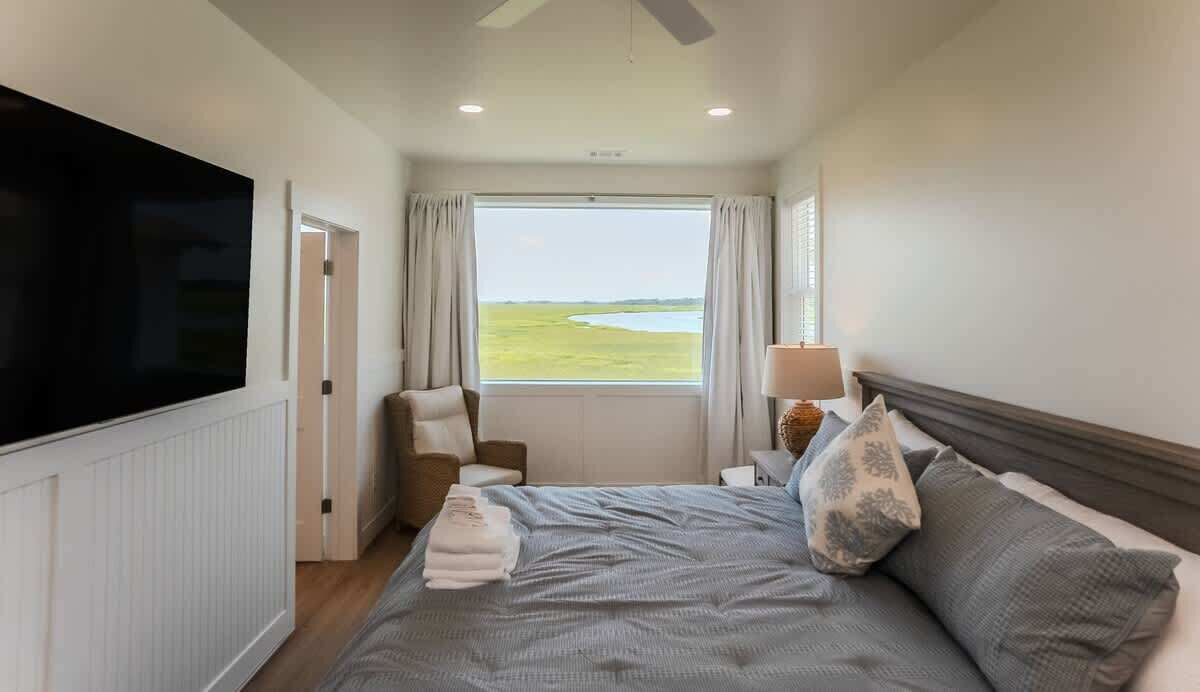 A hotel room with a bed, a TV, a window with a view of a field, a chair, and a lamp.