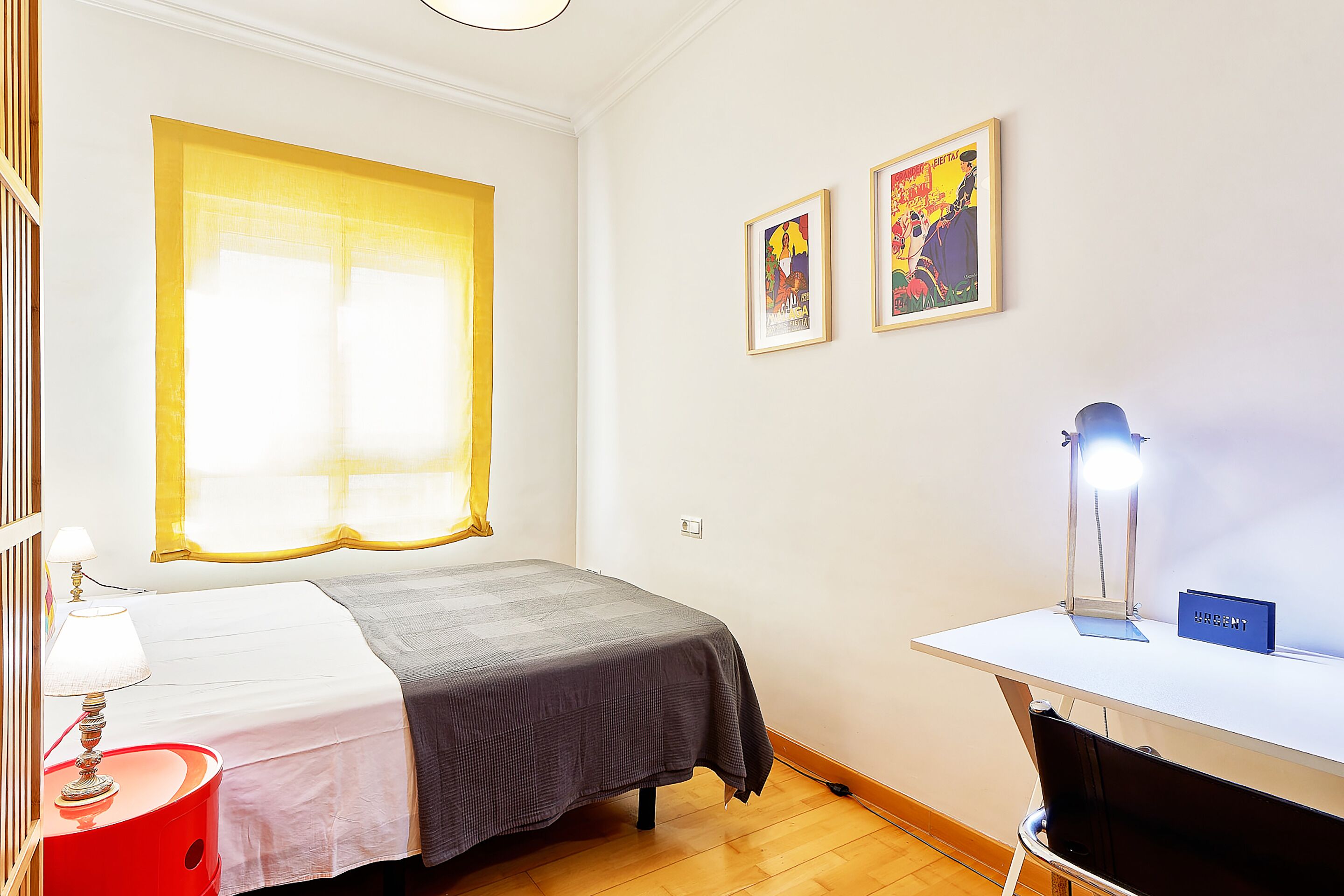 A room with a bed, a desk with a lamp, and two framed pictures on the wall.