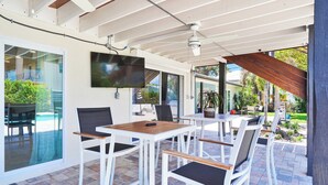 Un patio cubierto con mesas y sillas blancas, un televisor de pantalla plana montado en la pared y vistas a una piscina y follaje al exterior.