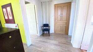 Un couloir avec un plancher en bois, une chaise bleue, une porte en bois et un mur jaune orné d’un tableau encadré.