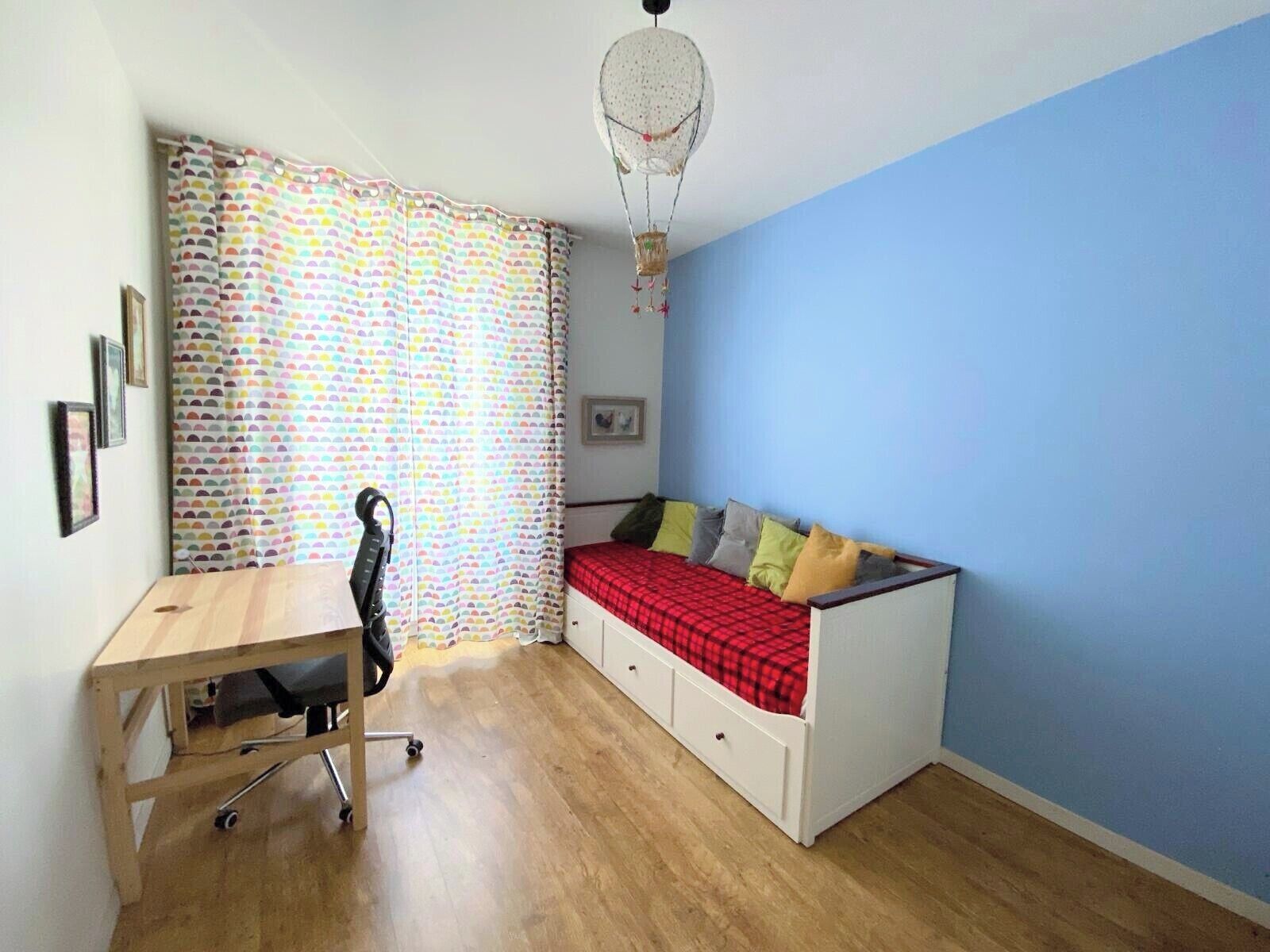 A small room with a desk, a bed with pillows, a colorful curtain, and a hanging light fixture.