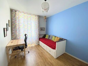 A small room with a desk, a bed with pillows, a colorful curtain, and a hanging light fixture.