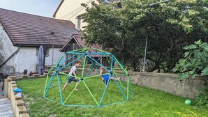 Un jardin à l’arrière d’une maison, avec une structure de jeu en métal vert, une terrasse en bois et une maison dont le toit est recouvert de tuiles.