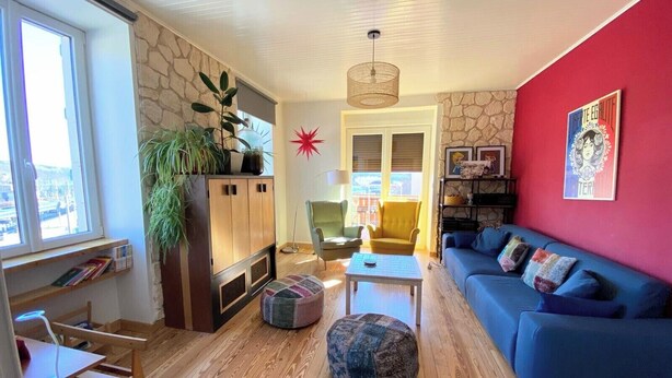Un salon avec un canapé bleu, un meuble en bois, un petit pouf rond et un mur rouge orné d’une affiche.