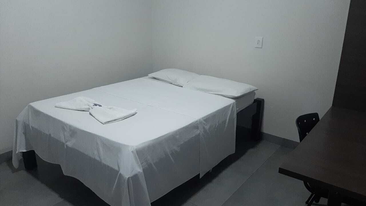 A single bed with white linens and a folded towel, a small black chair, and a wooden table in a simple room with plain walls and a tiled floor.