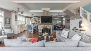 A spacious living room with a stone fireplace, a large grey sofa, and a dining area with a high ceiling. - Charming 3-bedroom house steps to Pine Point Beach with backyard oasis (Scarborough)