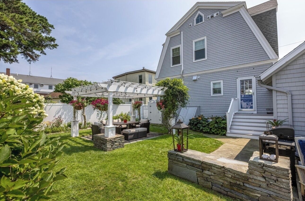 A residential backyard with a pergola, outdoor seating, a barbecue area, and a stone retaining wall.