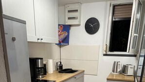 A compact kitchen with a refrigerator, stove, oven, and a small sink. There is a clock on the wall and a window with blinds. - Bungalow on Lake Jersleber See near Magdeburg (Barleben)