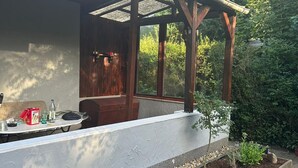 A covered outdoor seating area with a wooden pergola, a table with various items, and a garden with plants and a tree stump. - Bungalow on Lake Jersleber See near Magdeburg (Barleben)