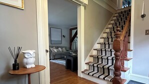 Un escalier recouvert d’une moquette à rayures, une rampe en bois et une porte blanche donnant sur un salon avec un canapé gris et une table basse.