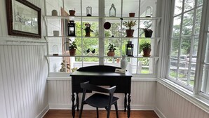 Une pièce comprenant un bureau en bois, une chaise, une fenêtre donnant sur un paysage verdoyant, ainsi que des étagères ornées de plantes en pot et d’objets décoratifs.