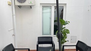 A patio area with two black chairs, a small table, and a potted plant. There is a white door and an air conditioning unit in the background.