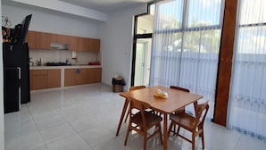 A modern kitchen with wooden cabinets, a dining area with a table and chairs, and large windows with sheer curtains. - Bonewood Guesthouse (Canggu)