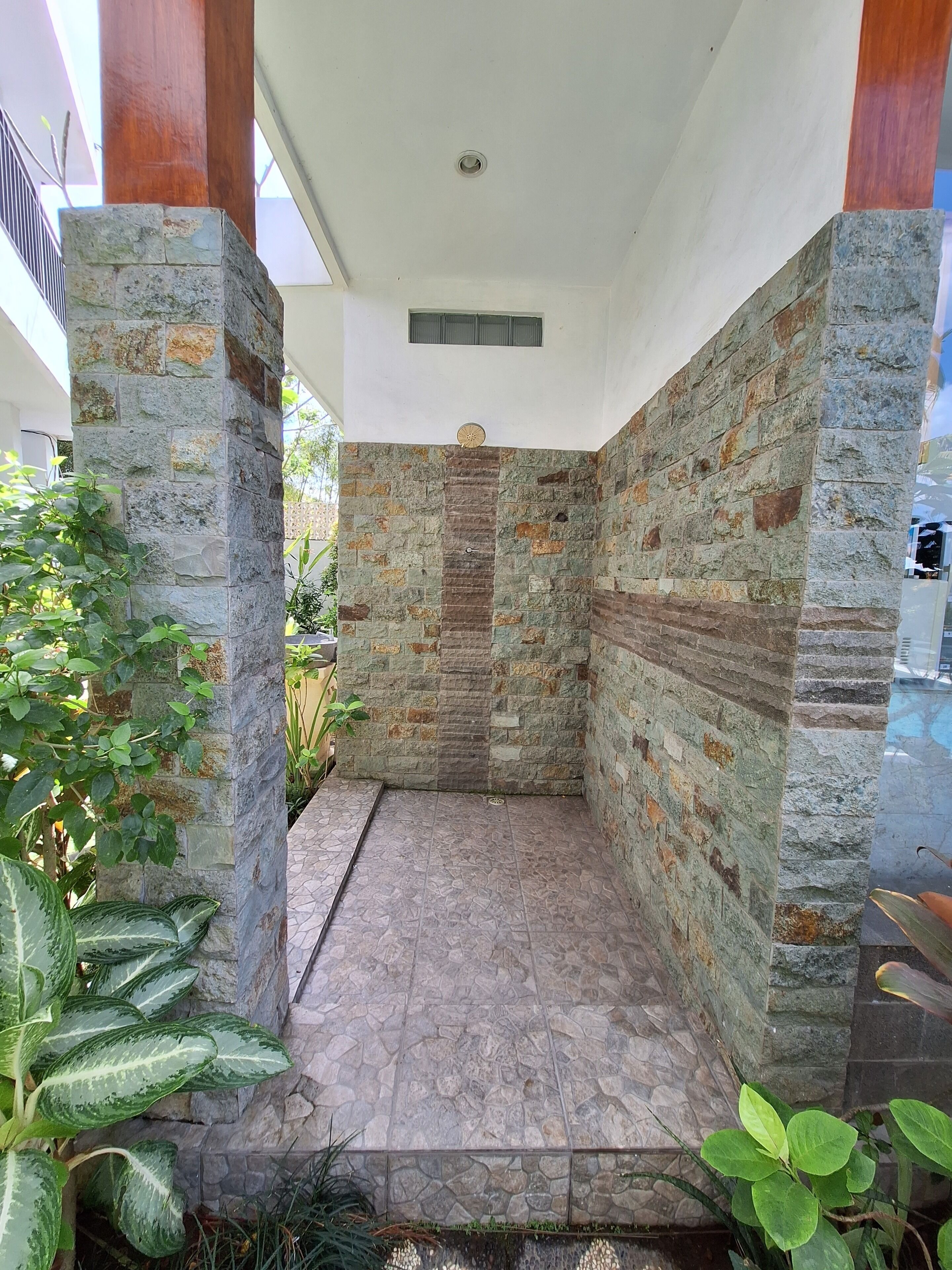 Un área de ducha con paredes de piedra y suelo de baldosas, rodeada de follaje.