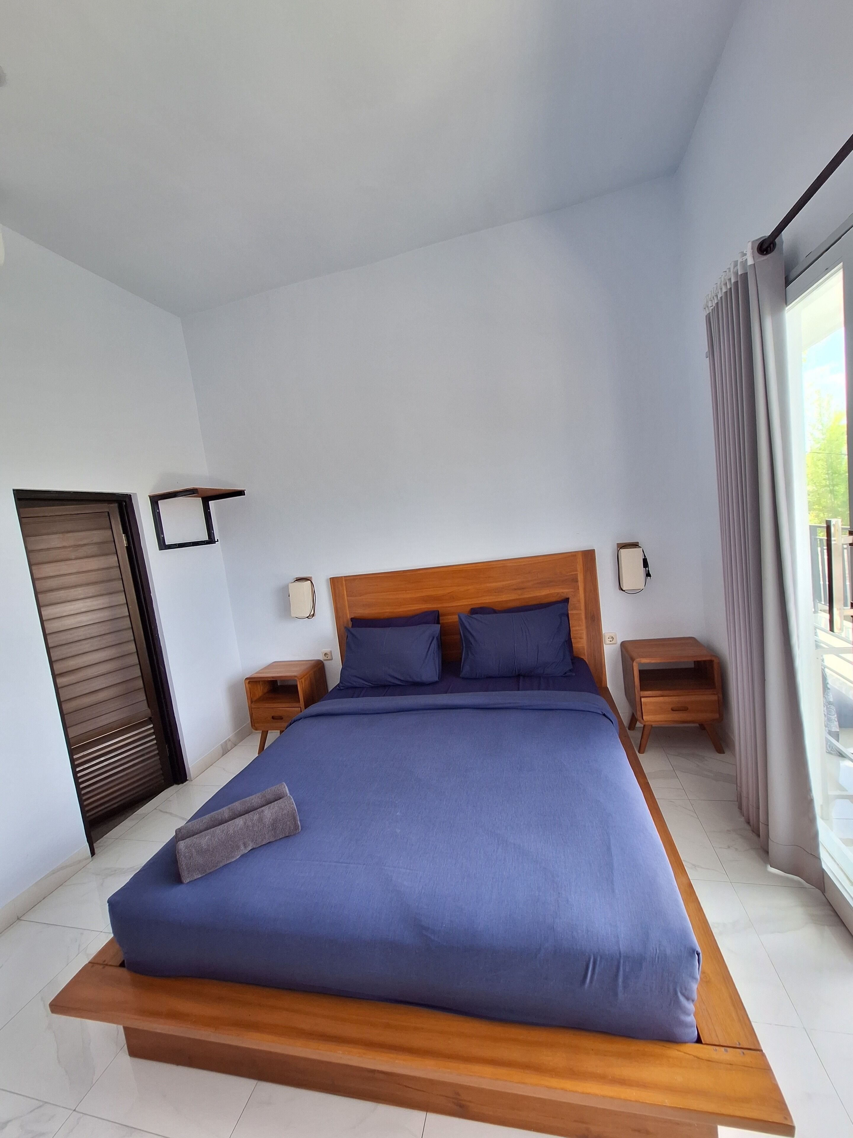 A modern bedroom with a large bed, wooden headboard, two bedside tables, and a view of the outdoors through a large window.