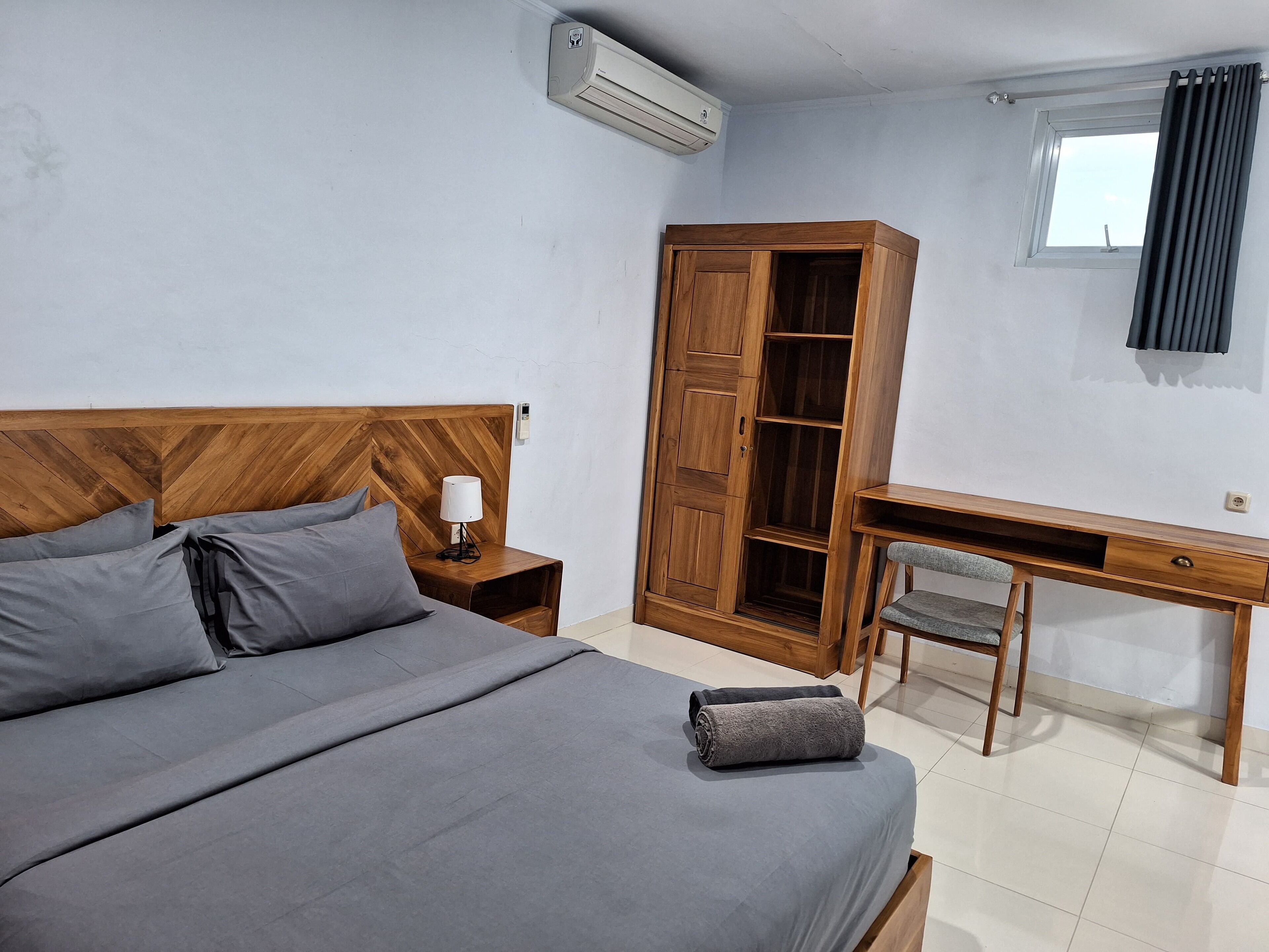 A bedroom with a wooden bed, a nightstand with a lamp, a wardrobe, a desk, and a chair.