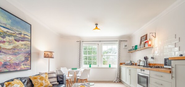 A modern kitchen with white cabinets, a gas stove, and a microwave. There is a dining area with a white table and chairs, and a large abstract painting on the wall. - Abbey Street (No 8) (St Andrews)