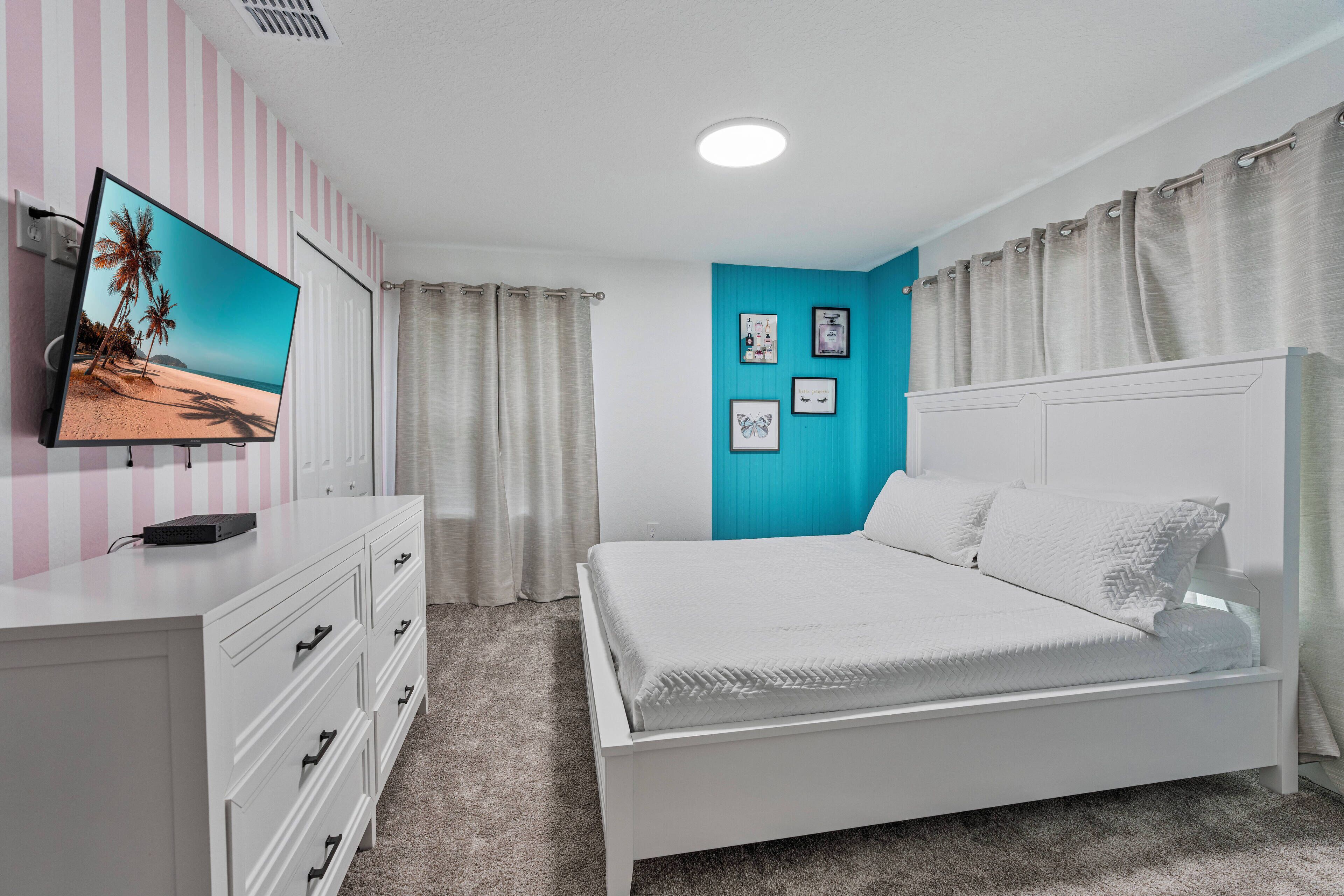 A bedroom with a bed, a TV on a dresser, a window with curtains, and wall art.