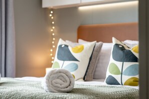 A neatly made bed with a rolled towel, pillows with a bird pattern, and a string of lights on the wall.
