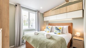 A neatly made bed with a green blanket, a wooden headboard, and decorative pillows. There is a small bedside table with a lamp, and a window with curtains. - Lodge No 83 | Hot Tub | Stewart's Resort Cameron (Cameron)