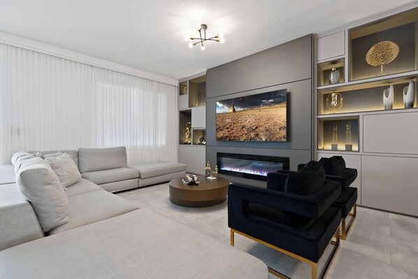 Una sala de estar moderna con un sofá seccional, un sillón oscuro, una mesa de centro redonda y un televisor de pantalla plana montado en la pared.