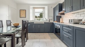 A modern kitchen with dark cabinets, a glass dining table, and a window providing natural light. - Kinness Place (No 6) (St Andrews)