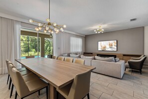 A modern living room with a wooden dining table, beige sofas, and a large window with curtains.