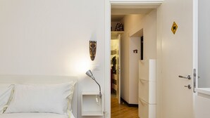 Een hotelkamer met een bed, een nachtkastje met lamp, een kledingkast en een deur naar een hal met kapstok en een koelkast.