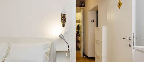 A hotel room with a bed, a bedside table with a lamp, a wardrobe, and a door leading to a hallway with a coat rack and a refrigerator.