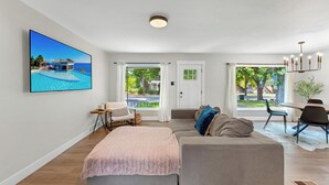 A spacious living room with a large flat-screen TV displaying a tropical resort, a sofa, a dining area, and a view of the outdoors. - Relaxing & Stylish: Close to Downtown ~ Yard ~ Pkg (Salt Lake City)